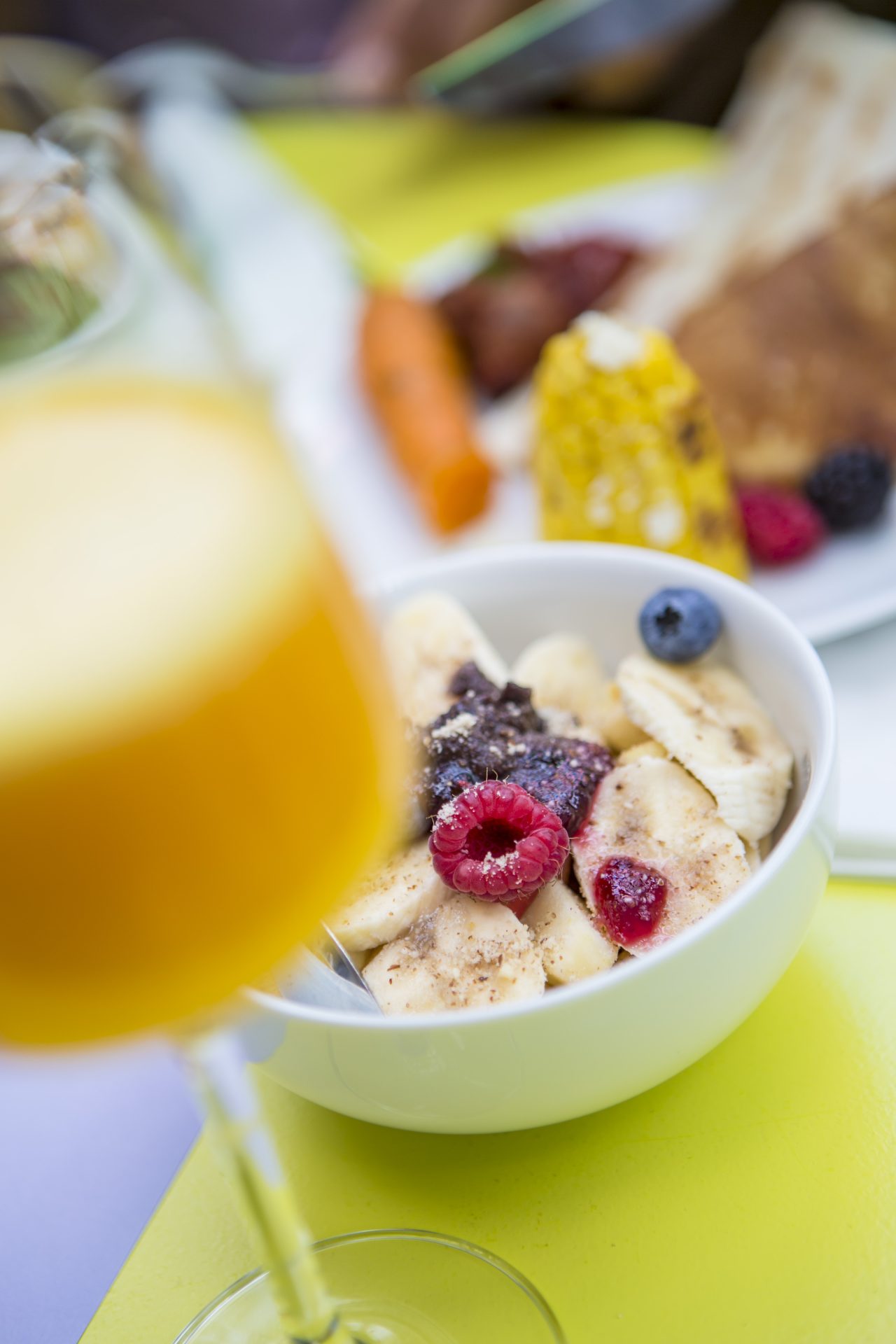 Bowl with bananas, raspberries, and blueberries, with a glass of orange juice in the foreground.