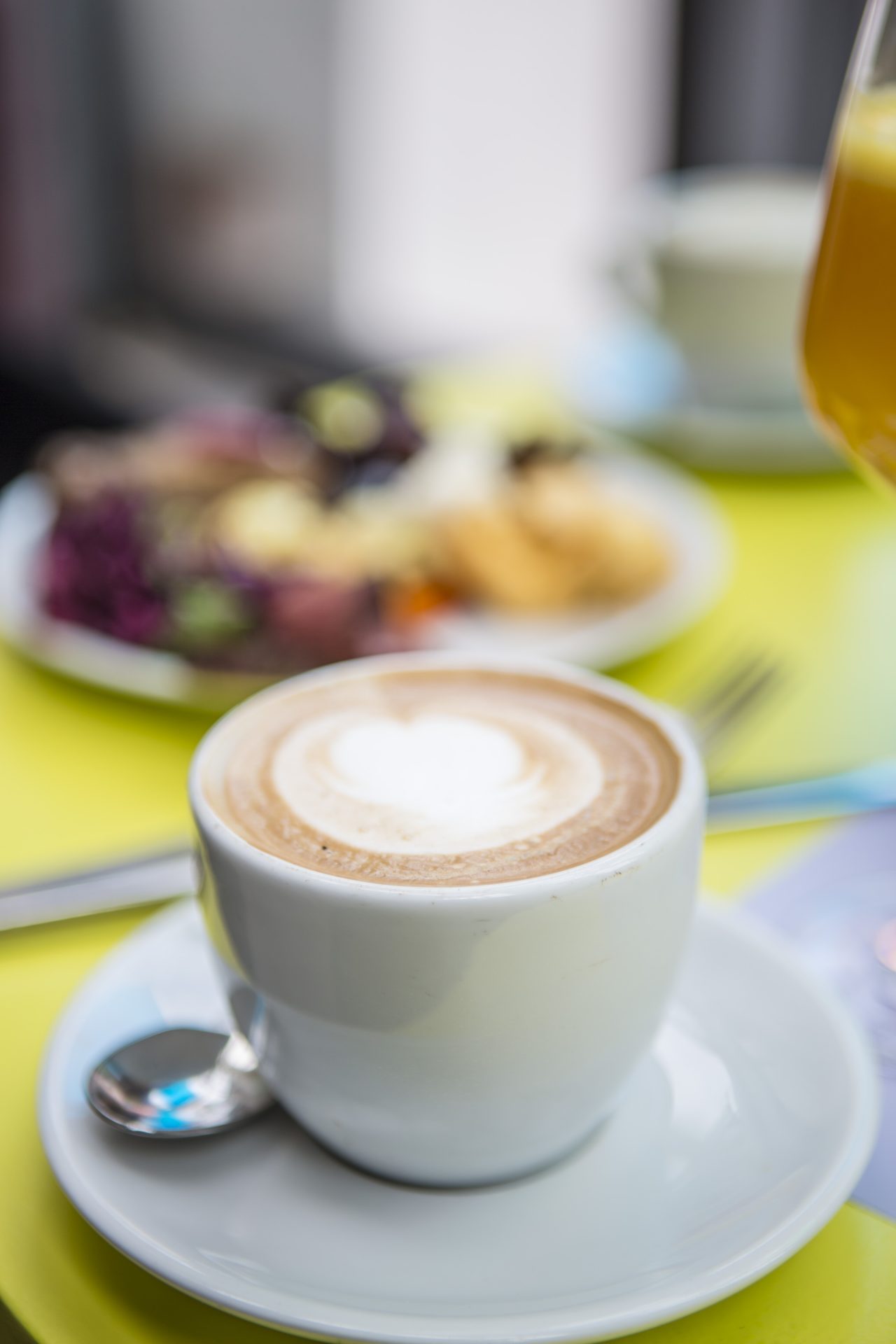 Coffee cup with milk foam, colorful plate with fruits and a drink in the background on a yellow table.