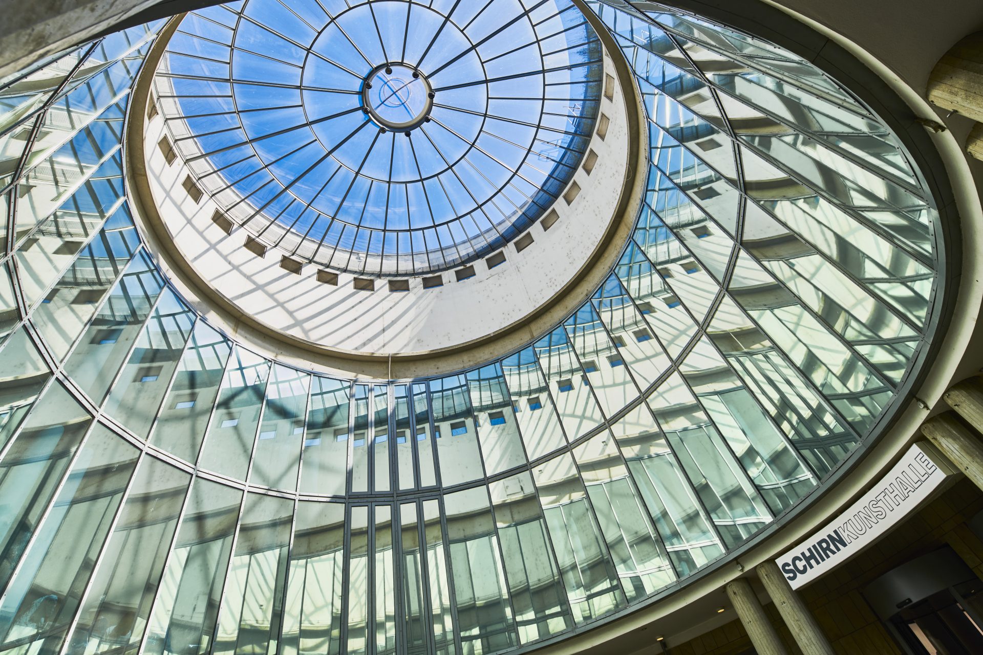Kreisförmige gläserne Kuppel mit blauem Himmel, lichtdurchlässig, Sicht von unten nach oben.