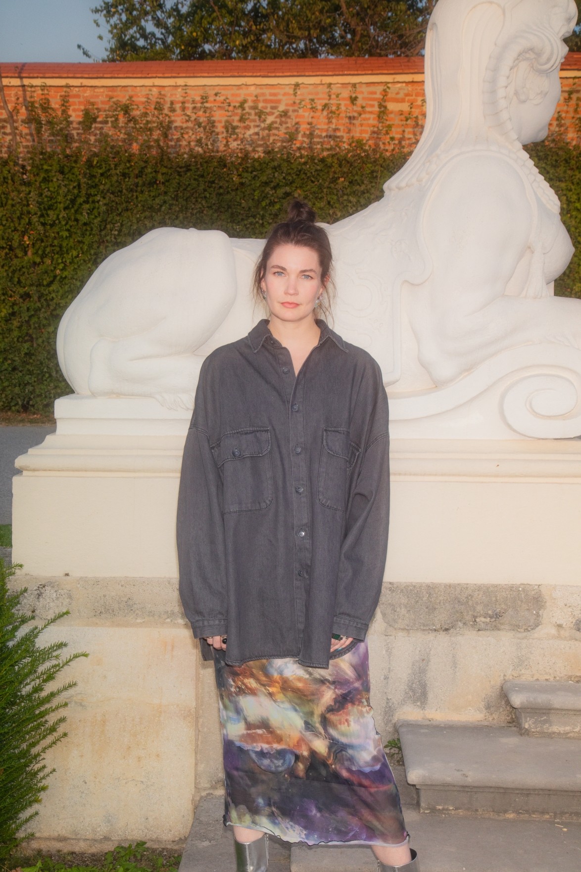 Annekathrin Kohout can be seen wearing a grey shirt and a colorful skirt. She is standing in front of a large white sculpture.